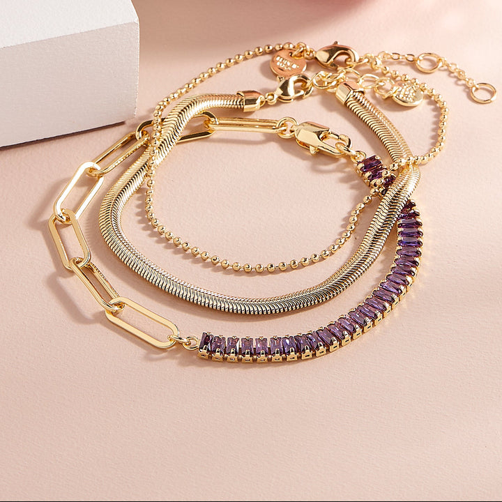 Set of three stacked bracelets on a ceramic plate — a crystal tennis bracelet, a bead bracelet with a rectangular blue birthstone, and a chain bracelet.