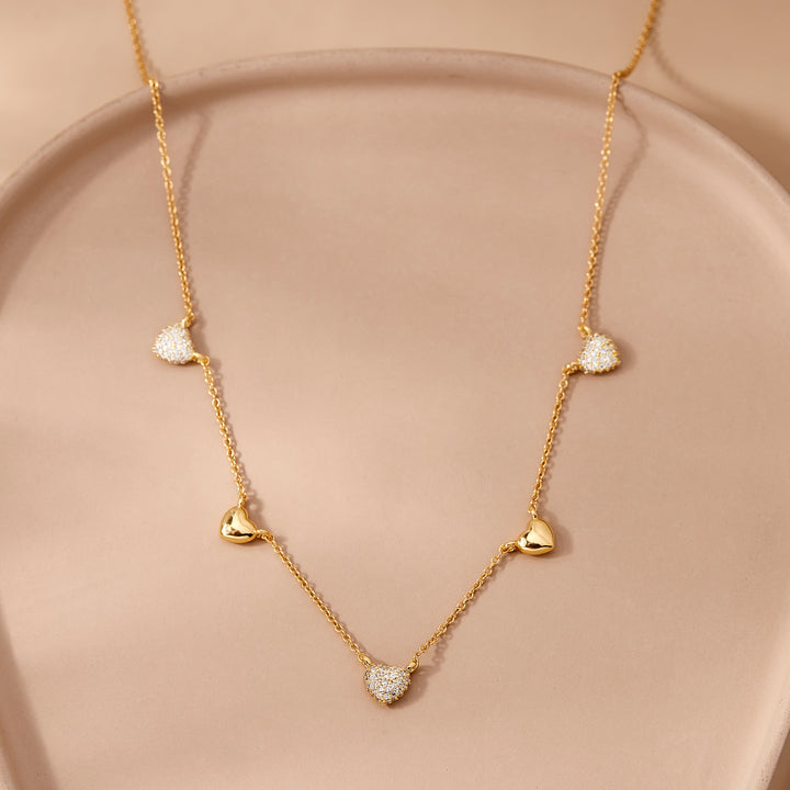  Posy Crystal Heart Necklace draped on a ceramic dish, with five heart charms alternating between pavé crystal and polished styles.