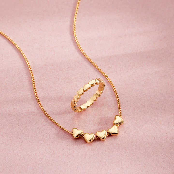  Posy Heart Necklace with five polished puffy hearts on a chain, styled alongside a matching heart eternity ring on a pink surface.