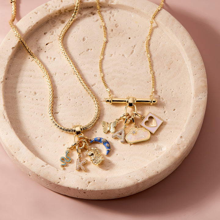 Poppy Charm Necklace styled with mixed charms including a blue moon, snake, initial, and bear, alongside a second charm necklace on a stone tray.