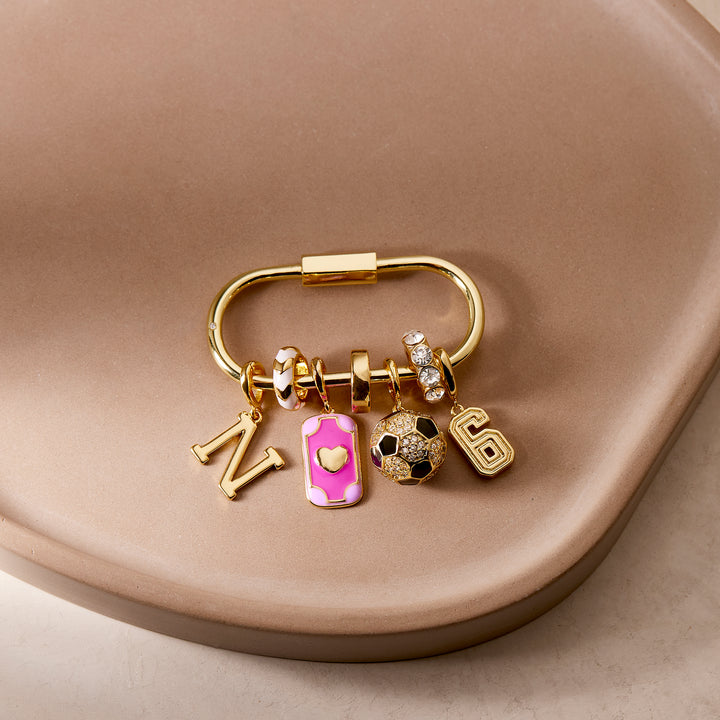 Oval carabiner-clasp bag charm key chain styled with an initial charm, pink enamel rectangular charm, soccer ball charm, and number charm on a ceramic dish.