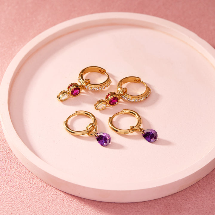 Two styles of birthstone huggie hoop earrings on a tray, one pair with pavé hoops and stacked stone charms, another with purple teardrop stone drops.