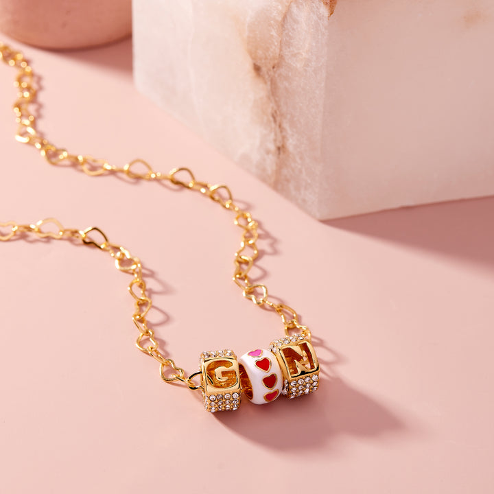  Initial letter charm necklace with crystal-edged cubic charms and a white enamel heart bead, on a heart-link chain, close-up flat lay.