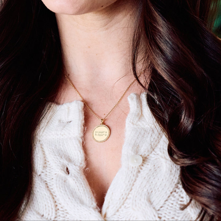 Cece Coordinate Necklace with a round pavé-bordered disc pendant engraved with custom coordinates, on a chain on a models neck.