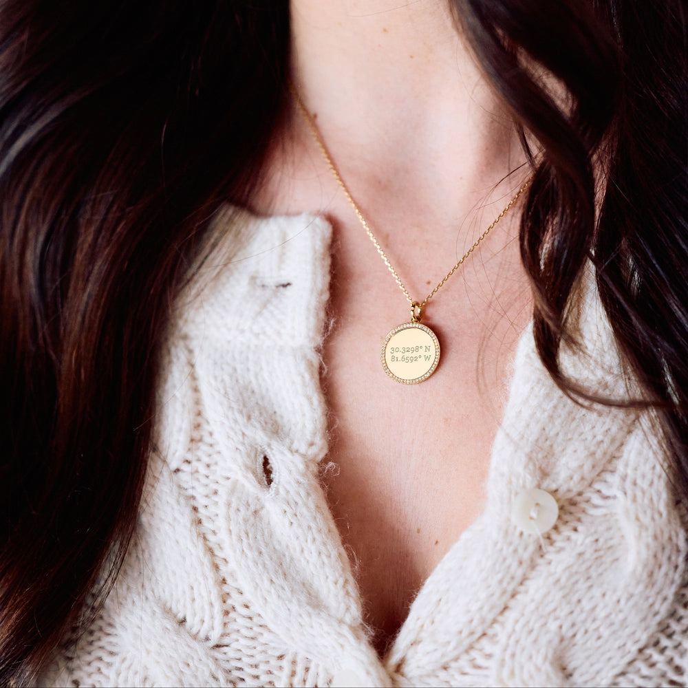  Cece Coordinate Necklace with a round pavé-bordered disc pendant engraved with custom coordinates, on a chain on a models neck.