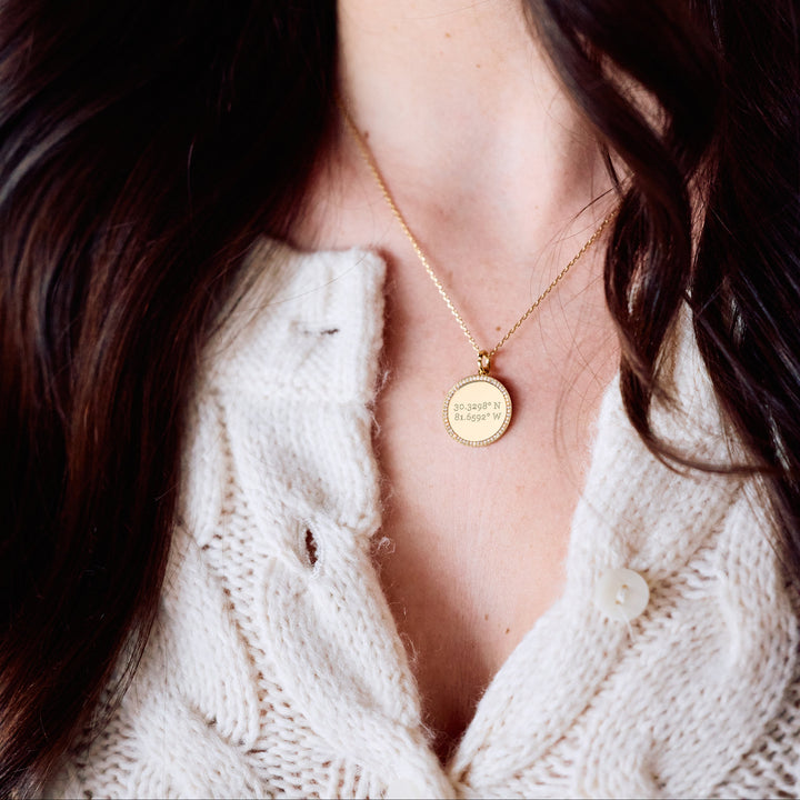 Cece Coordinate Necklace with a round pavé-bordered disc pendant engraved with custom coordinates, on a chain on a models neck.