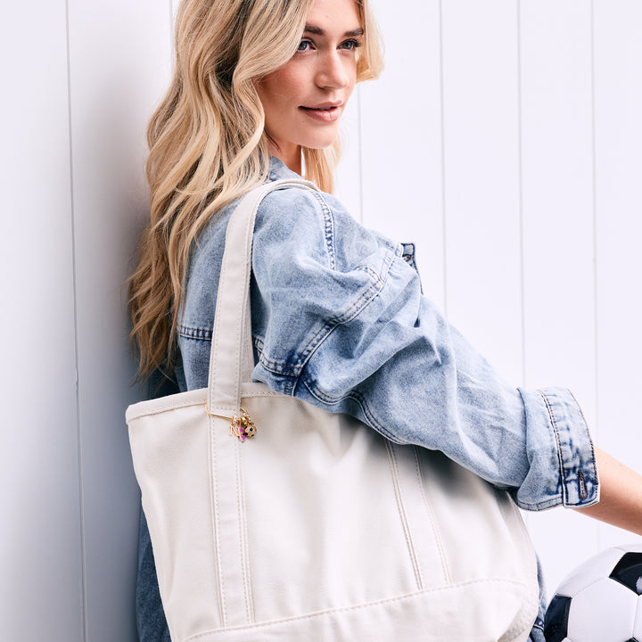 Charm key chain with an initial letter, pink enamel rectangular tag, crystal soccer ball, and number charm, clipped to a white canvas tote carried by a model in a denim jacket.