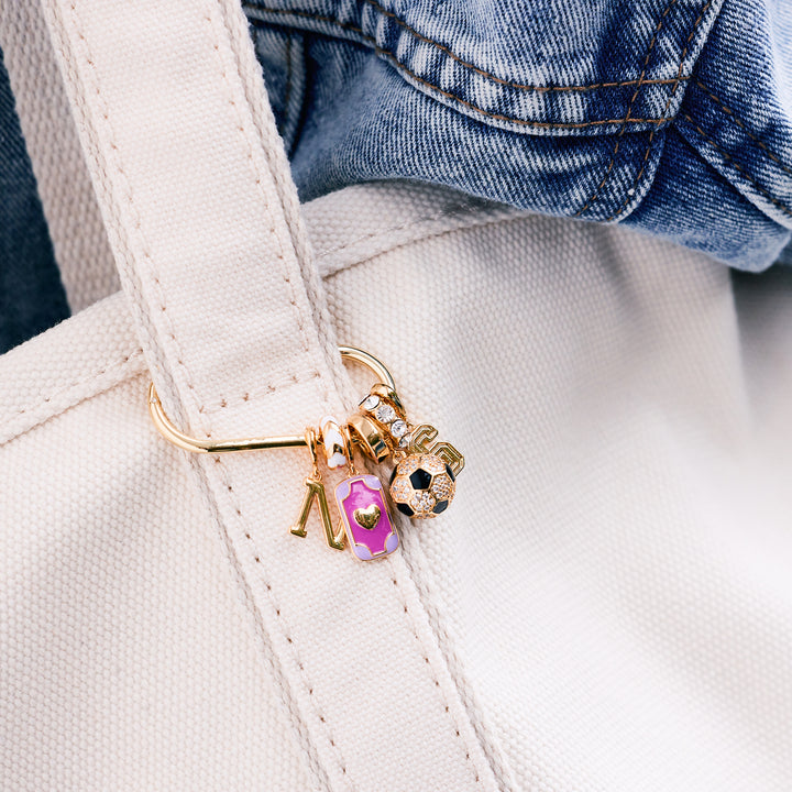 Charm key chain with an initial letter, pink enamel rectangular tag, crystal soccer ball, and number charm, clipped to a white canvas tote carried by a model in a denim jacket.
