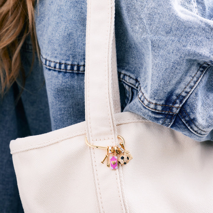 Charm key chain with an initial letter, pink enamel rectangular tag, crystal soccer ball, and number charm, clipped to a white canvas tote carried by a model in a denim jacket.
